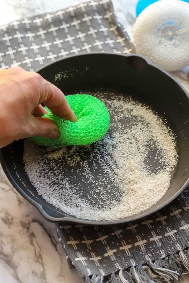 Green nylon scrub brush cleaning a cast iron pan with salt