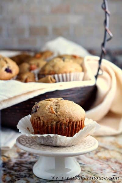 Single persimmon muffin on small cake stand with basket of muffins in background