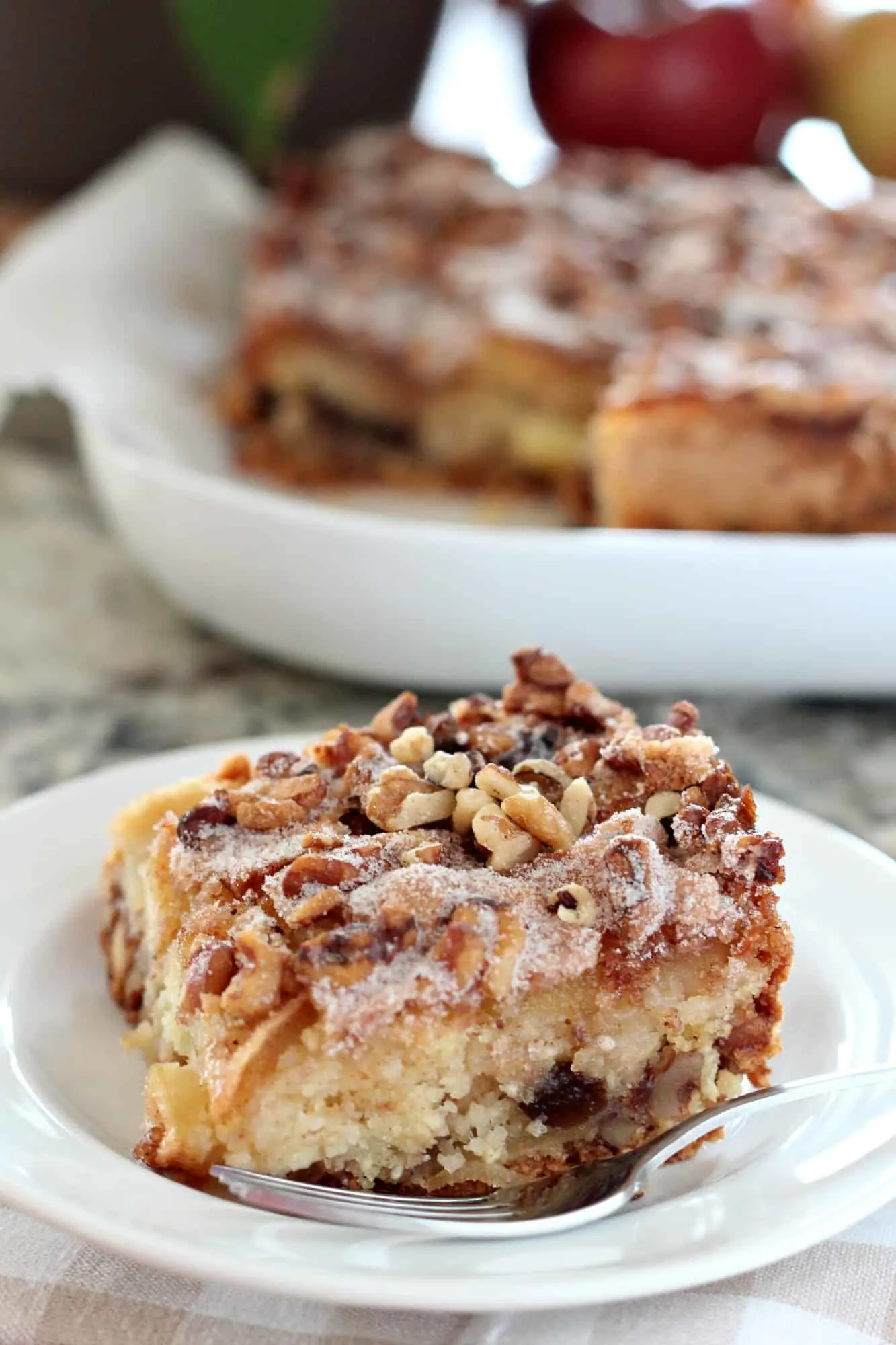 One square slice of Apple Cake for Passover on a white plate with the rest of the cake in the background