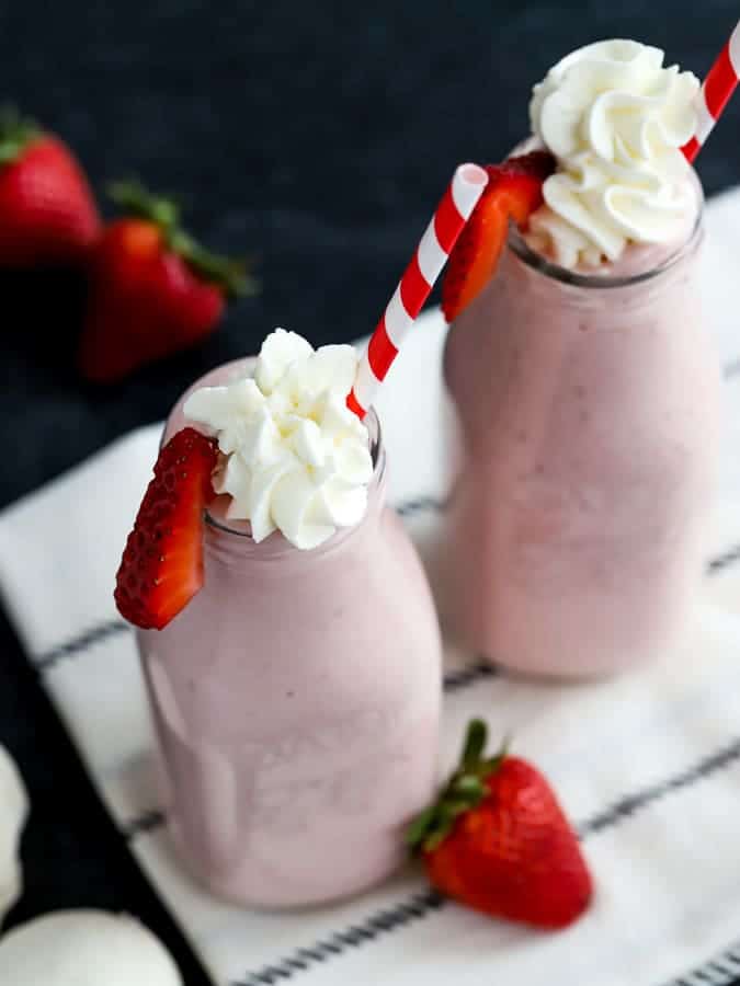 Two strawberry milkshakes in glass milk bottles with whipped cream and red and white striped straws