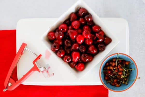 Oxo cherry pitter with pitted cherries in a white bowl and cherry pits in small blue bowl