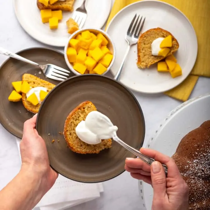 Mango bundt cake on gray plate with hands adding dollop of yogurt