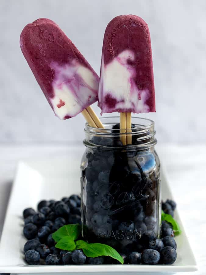 Two magenta colored blueberry cream popsicles in a mason jar filled with fresh blueberries