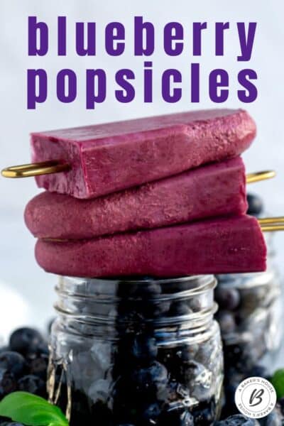 Three blueberry popsicles stacked on top of a small mason jar filled with blueberries