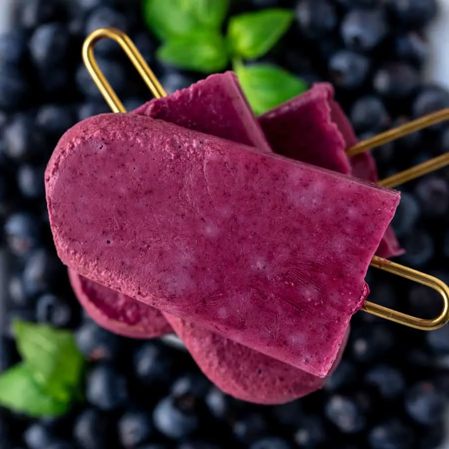 Frozen blueberry popsicles stacked in a group of three with gold handles on top of fresh berries