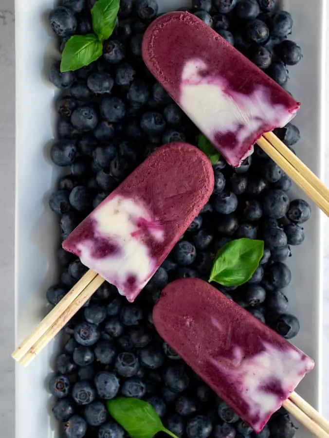 Three blueberry and Greek Yogurt vanilla creamsicles laying on top of fresh blueberries with basil