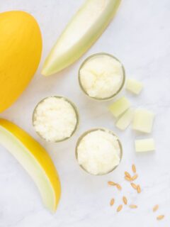 Overhead image of three mason jars of melon granita with canary melon and slices and seeds on marble surface