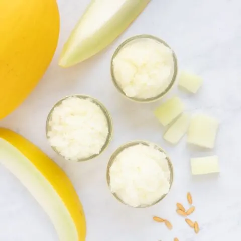 Overhead image of three mason jars of melon granita with canary melon and slices and seeds on marble surface