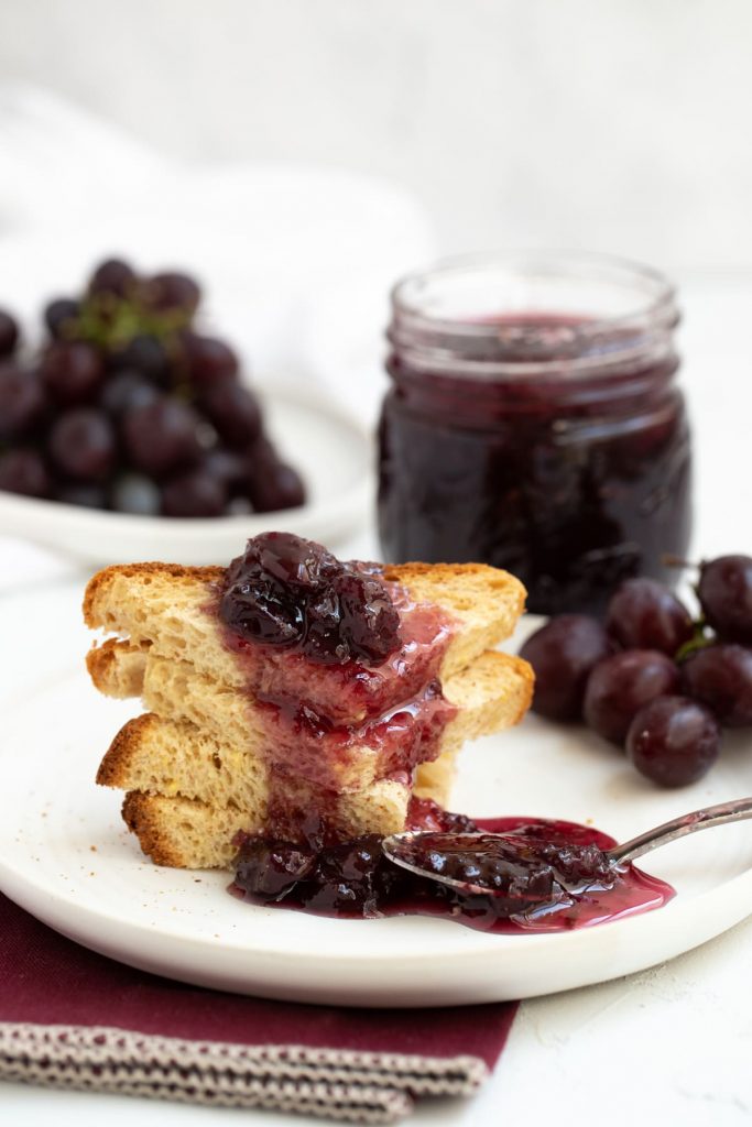 Four stacked quarters of toast with grape jam dripping over and mason jar of jam in background