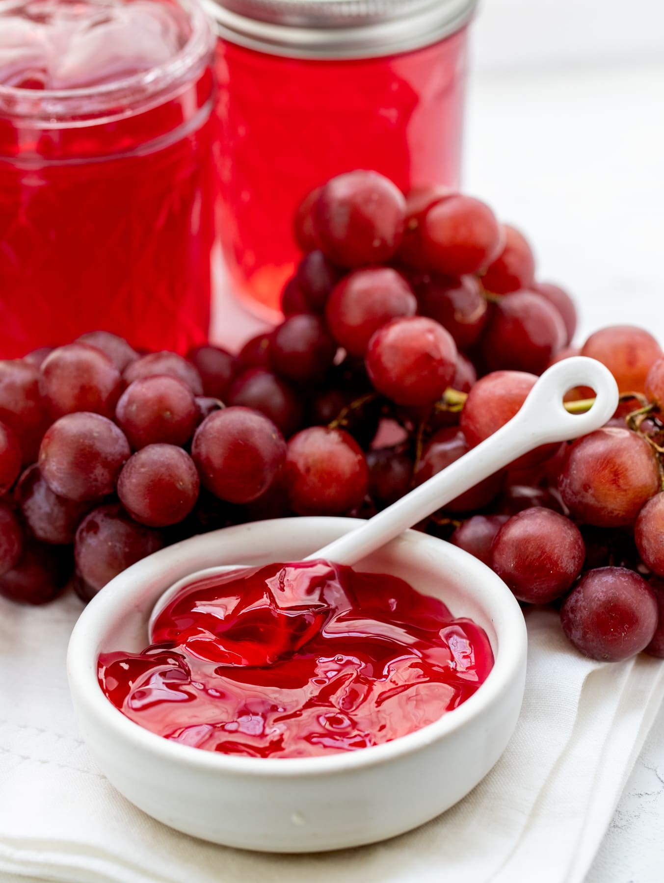 White bowl filled with grape jely with fresh grapes and jars of jelly in background