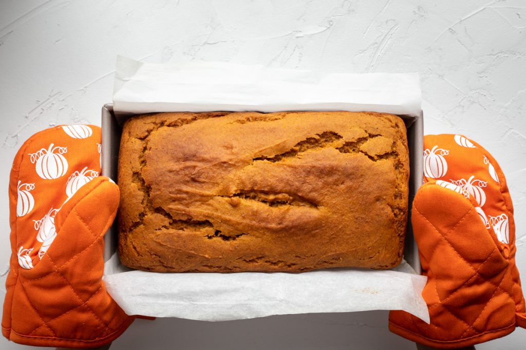 Freshly baked loaf of pumpkin bread in loaf pan held by two pumpkin orange baking mitts