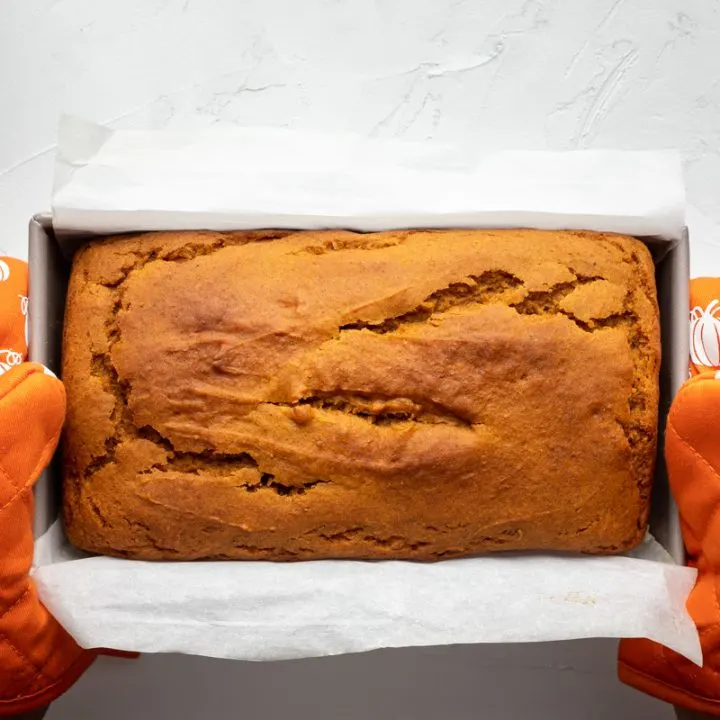 Fresh loaf of pumpkin bread held by two orange pumpkin baking mitts