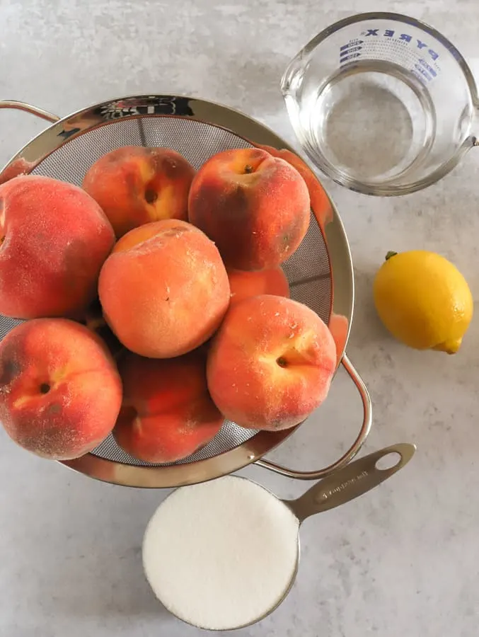 bowl of freshly picked peaches, cup of sugar, water, and a lemon on a gray surface