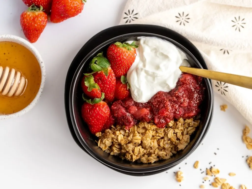 Black bowl with plain yogurt, whole strawberries, granola and jam