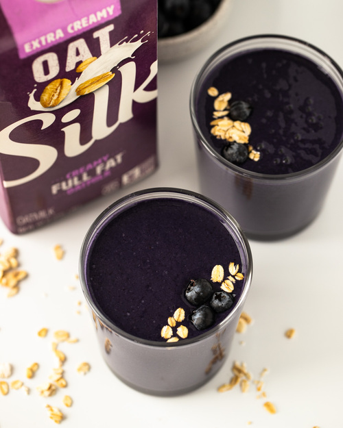 two glasses of blueberry smoothie next to carton of Silk Oatmilk