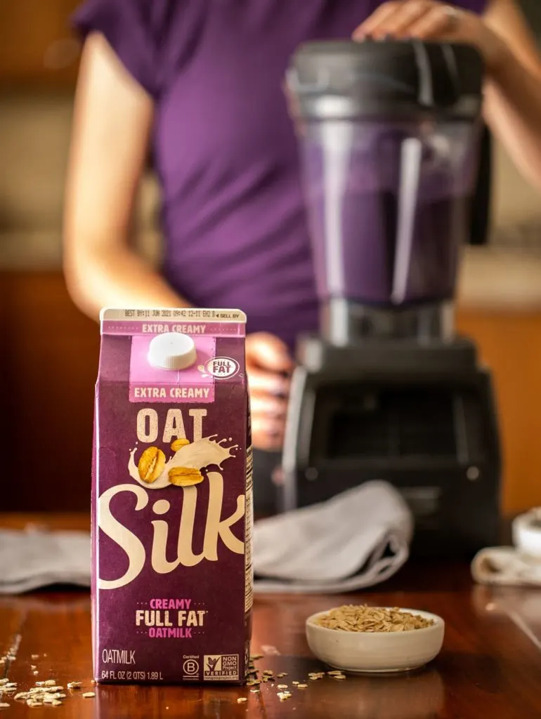 carton of Silk Oatmilk with person wearing purple shirt using blender to make blueberry smoothie
