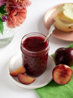 jar of plum butter on white pottery plate with fresh plums and flower vase