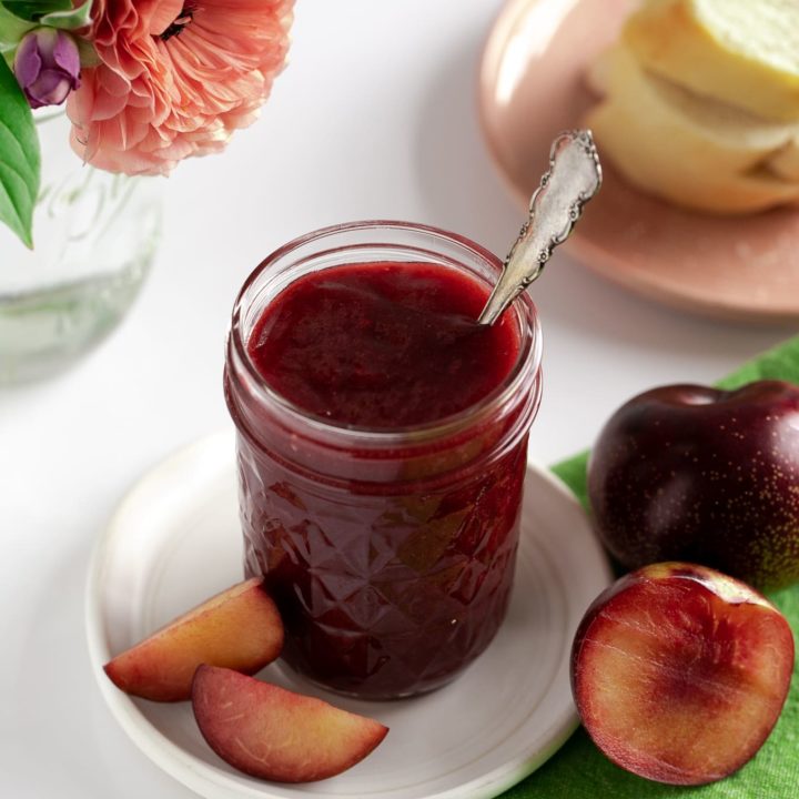 jar of plum butter on white pottery plate with fresh plums and flower vase