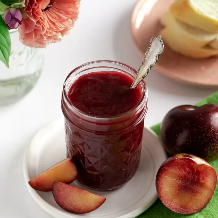 jar of plum butter on white pottery plate with fresh plums and flower vase